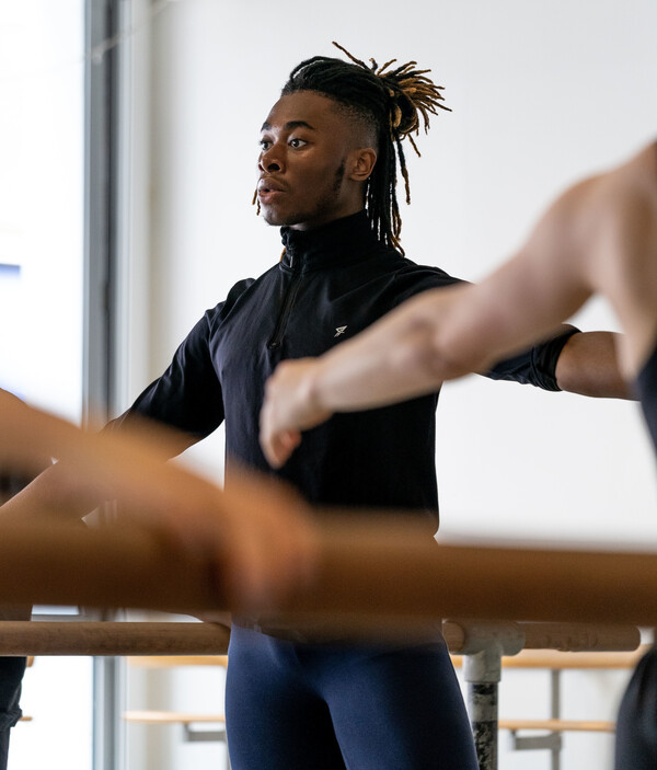A focused ballet dancer in black attire grips a barre, arm extended, in a Centre for Advanced Training studio, showcasing the dedication to classical ballet technique within the program.