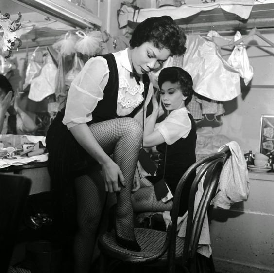 A behind-the-scenes shot of two chorus girls at the Forbidden City nightclub preparing for their performance, highlighting the anticipation and energy before taking the stage and showcasing the dedication of the performers.