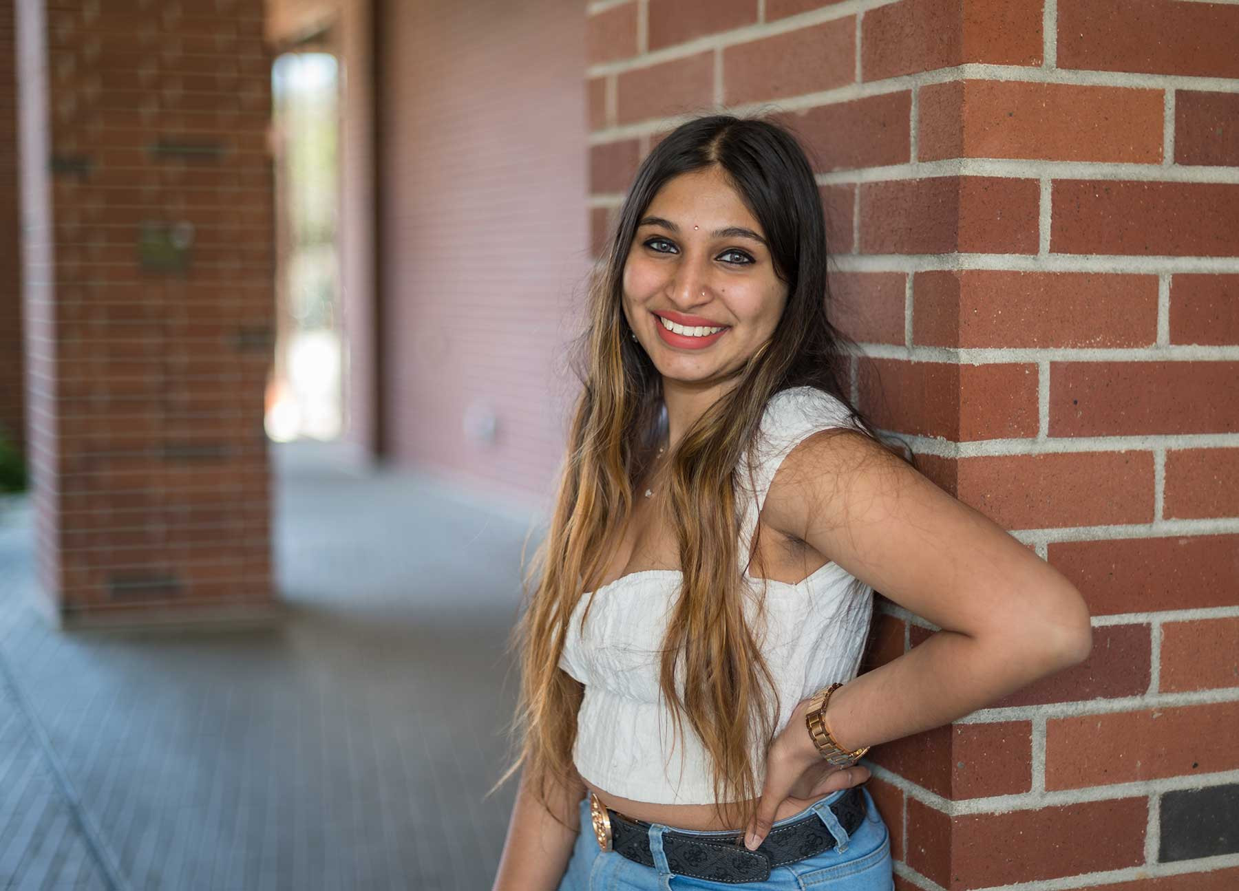 Aishwarya Gowda poses against a brick pillar on campus, wearing a white shirt and jeans.