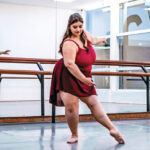 Júlia Del Blanco wears a burgundy camisole leotard and matching ballet skirt and pink ballet slippers. She is shown in a dance studio doing a tendu devant in effacé with her left leg extended and her right leg in plié.