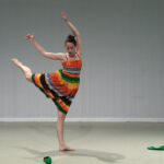 Monica Thomas in a multi-colored dress stands barefoot in attitude back, two green shoes scattered on the floor near her.